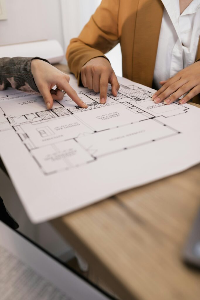 Close-up photograph of hands pointing to architectural plans printed on white paper. The plans show a floor layout with lines and symbols, suggesting a house design. The hands are gesturing, possibly indicating a discussion or explanation of the plans. The image illustrates the "Planning and Design" stage of Brownhill Homes' services, as described in the accompanying text, which focuses on understanding client needs and creating detailed designs.