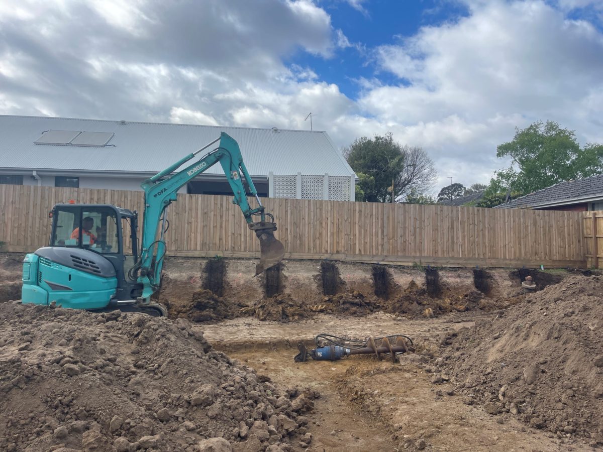 Photograph of a construction site with an excavator digging into the ground. The excavator is teal and black, and the ground shows dug-up earth and a trench. In the background, there is a fence and some trees. The image represents the "Site Preparation and Construction" stage of Brownhill Homes' services, as described in the accompanying text, which focuses on preparing the site for building and the construction process itself.