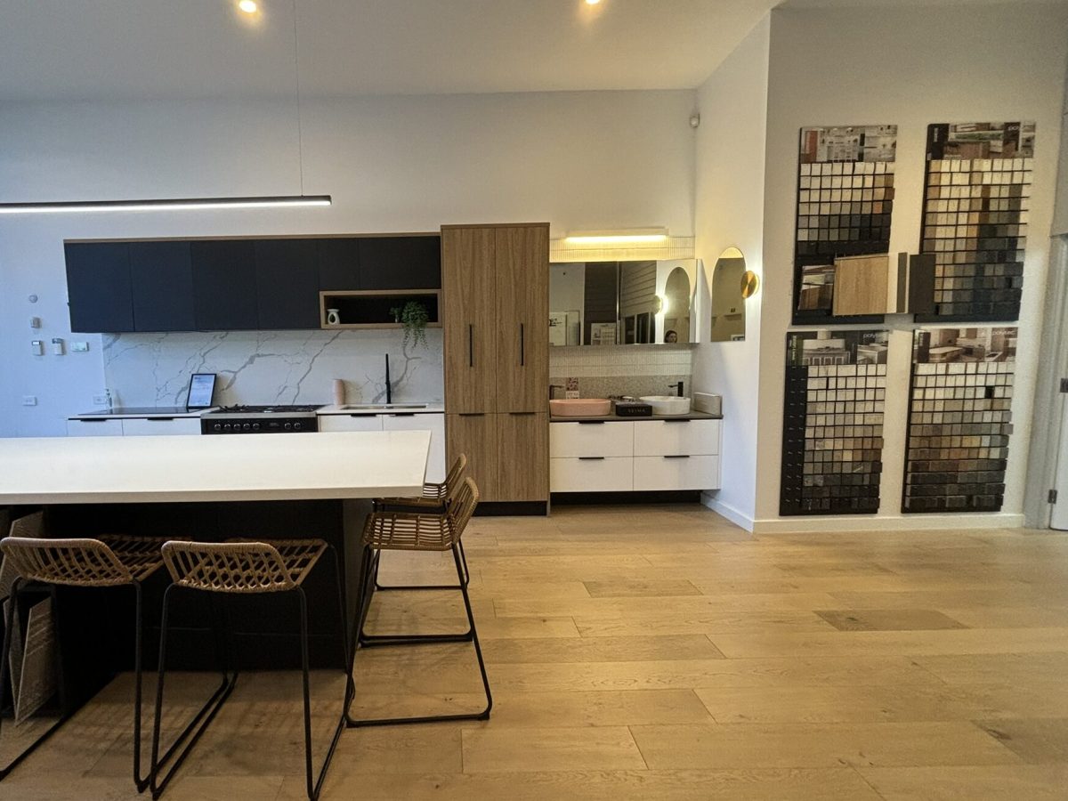 Interior photograph of a modern showroom with a kitchen display. The kitchen features a white countertop island with two woven bar stools, dark cabinetry on the left wall, and a light-colored cabinet unit with a mirror on the right. Framed artwork is visible on the right wall. The showroom has light-colored flooring and walls, creating a bright and contemporary atmosphere. The image is used to represent Brownhill Homes' "Modern Showroom," as described in the accompanying text, which focuses on exploring design possibilities and visualizing future homes.