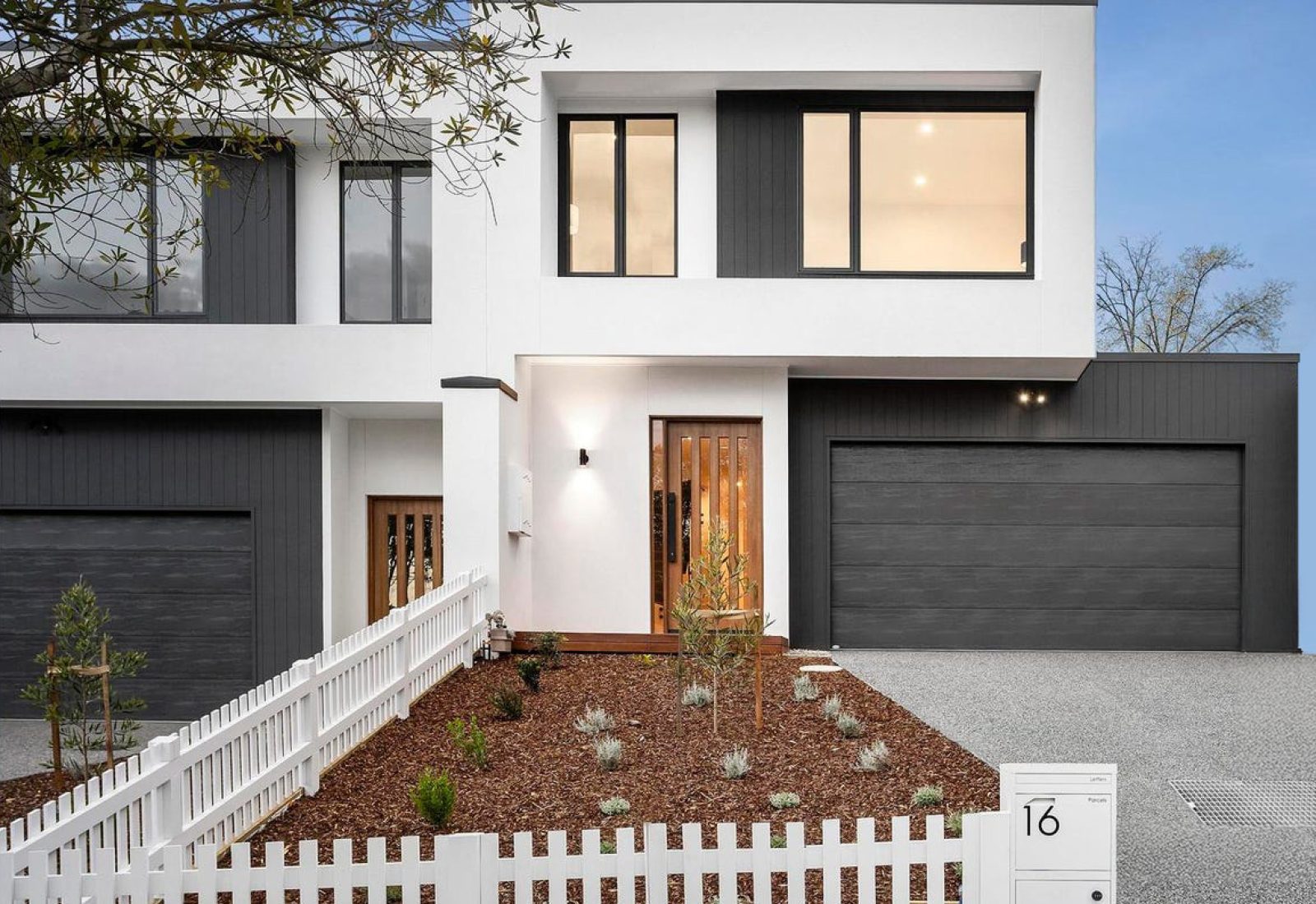 Exterior photograph of a modern, two-story house with a white facade and dark gray garage doors. The house features rectangular windows, a dark front door, and a small garden with brown mulch and young plants. A white picket fence runs along the front of the property, with a house number "16" visible on a small sign. The sky is blue with some light clouds. The image is used to represent Brownhill Homes' "Follow The Build" service, as described in the accompanying text, which focuses on sharing exclusive updates and behind-the-scenes insights during the building process. The address "16 Pentlowe Avenue, Templestowe Lower" is likely associated with this property, based on the AI model's information.