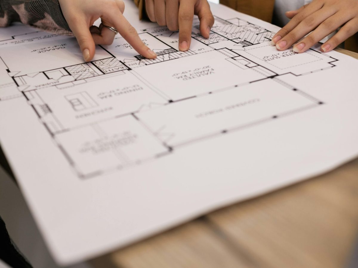 Close-up photograph of hands pointing to architectural plans printed on white paper. The plans show a floor layout with lines and symbols, suggesting a house design. The hands are gesturing, possibly indicating a discussion or explanation of the plans. The image illustrates the "Planning and Design" stage of Brownhill Homes' services, as described in the accompanying text, which focuses on understanding client needs and creating detailed designs.