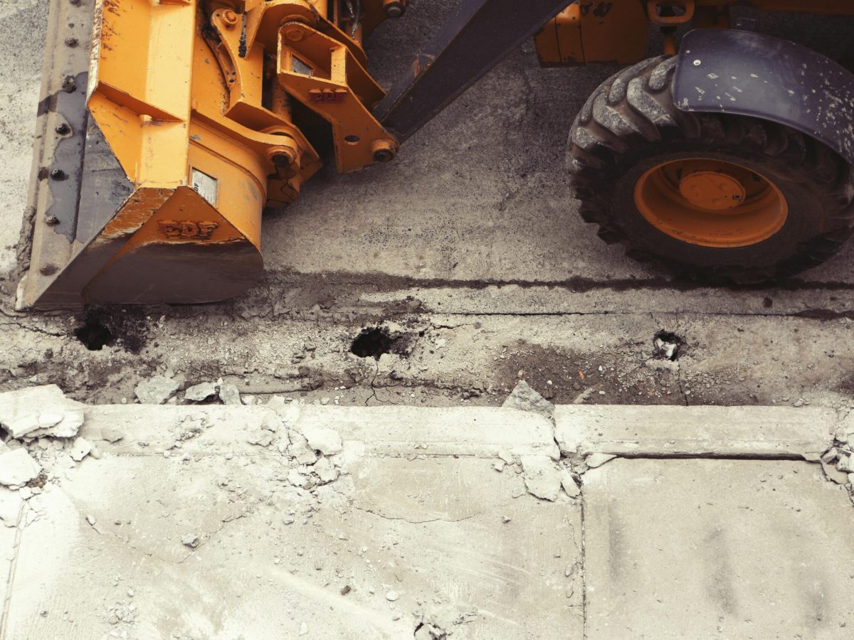 Close-up photograph of a construction vehicle with a large, orange arm and a black tire. The vehicle is positioned on a cracked concrete surface, suggesting demolition work. The arm and tire are the main focus of the image, with the rest of the vehicle partially visible. The scene represents the "Demolition" stage of Brownhill Homes' services, as described in the accompanying text, which focuses on managing the demolition process for a seamless build.