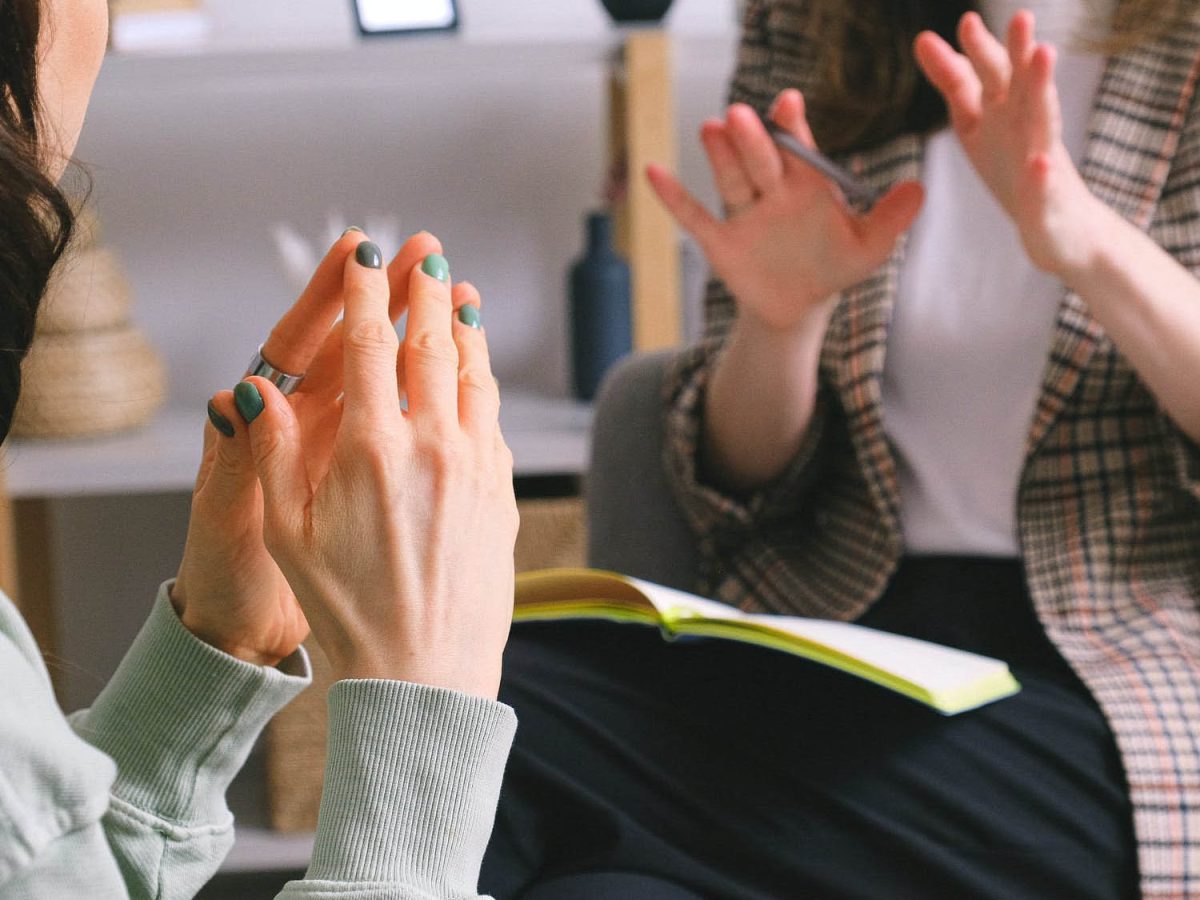 Two women are seated and talking, with one woman gesturing with her hands towards the other. A book is open between them, and a blurry object is visible in the background.