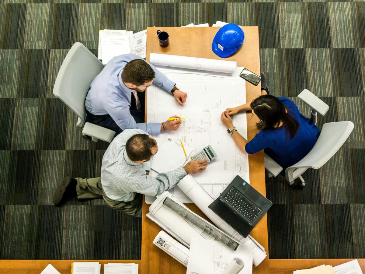 Overhead photograph of three people working around a large table covered with architectural blueprints. The people are dressed in business casual attire and are discussing the plans. A blue hard hat, a phone, and a laptop are also on the table. The image represents Brownhill Homes' "Project Tracking Made Easy" service, as described in the accompanying text, which focuses on providing an intuitive online portal for clients to stay informed throughout the building process.
