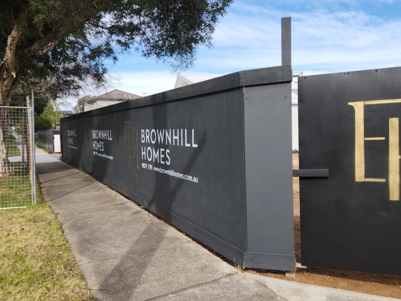 Exterior photograph of a construction site with a dark gray temporary fence. The fence has the "BROWNHILL HOMES" logo in white letters, along with website and phone information. The fence runs along a sidewalk with trees and houses visible in the background. The image represents the "demolition" stage of Brownhill Homes' knockdown rebuild process, as described in the accompanying text, which focuses on guiding clients from demolition to construction.